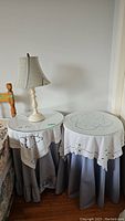 Two round side tables each with glass top and decorative white tablecloth over gray skirts, bedside lamp on one table