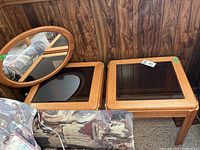 Two wood framed side tables with smoked beveled glass tops placed side by side next to a wood paneled wall, accompanied by an oval wood framed mirror.