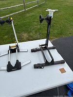Two foldable portable bike stands set on a white folding table outdoors with grass and a wooden fence in background. One silver Topeak stand and one black Feedback Sports stand.