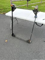 Full view of the black metal rectangular frame with two wheels and height adjustment mechanisms shown on a white folding table outdoors.