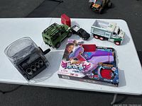 Wide view of the lot items on a white table outdoors: coin sorter, green toy truck, black small car, white and green garbage truck, and Nerf Rebelle Mega Twisted toy gun in packaging