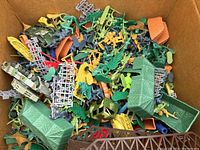 Box filled with assorted plastic toy soldiers and military accessories visible from above. Shows color variety and different items including vehicles and structures.