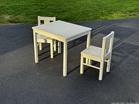 Photo of white kids table with two chairs outdoors on pavement, showing general condition and size relative to grass background.