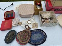 Collection of dollhouse braided rugs, fabric boxes, miniature porcelain bowl, tiny clock, folded newspapers, and assorted fabric pieces on a table.