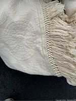 Close-up of cream-colored embroidered bedspread showing detailed quilted floral pattern and long fringed edges.