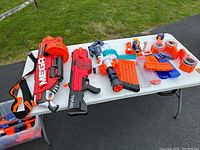Photo showing three large Nerf blasters including a Mega series blaster, Rival series blaster, and a white and orange blaster with multiple dart magazines and attachments.