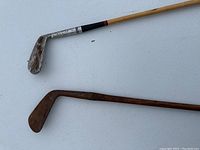 Two old golf clubs laid on a white surface showing the metal 9 iron head and the fully wooden club head.