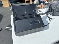 Fujitsu ScanSnap iX500 document scanner closed and placed on a white table outdoors, power and USB cables visible.