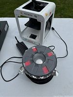 Toybox 3D printer with a power cord and a spool of filament in foil packaging on a table.