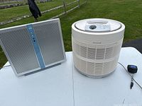 Two air purifiers on a white table outside: one rectangular silver and blue, one white cylindrical Honeywell model.