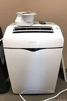 White Noma portable air conditioner with vent and remote on top, front view showing brand logo and missing exhaust tube attachment.