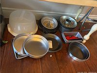 Image showing a variety of metal baking pans including round, square, and bundt cake pans alongside a white plastic cake storage container with lid, and a red oven mitt.