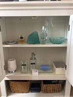 Full view inside bathroom cabinet showing multiple vanity items including perfume bottle, spray bottle, glass decorative item, soap dispenser, toothbrush holder, waste bin, soap bars, and woven baskets