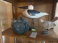 White standing bird with black and white pattern and metal legs, next to blue ceramic prone bird on wooden shelf