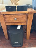 Subwoofer under small wooden table with two satellite speakers placed on top with wires behind.