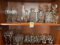 Shelf showing four clear glass decanters with metal tags, one clear glass pitcher, beer glasses, shot glasses, and other drinking glasses.