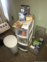 Overall view of plastic drawer unit with assorted light bulbs on and inside drawers, white step trash bin and plastic storage bin with light bulbs nearby.