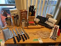 Wide shot showing the full kitchen tool lot including cutting knives, grater, rolling pin, cutting board, oil jars, measuring bowl, knife block, and food saver.