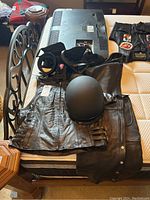 Motorcycle apparel items including black leather vest, chaps, gloves, and belt, laid out on bed with television in background (TV not included).