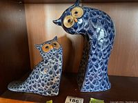 Front view of two owl ceramic bookends on wooden shelf, displaying size difference and textured blue pattern with yellow eyes.