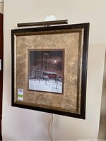 Framed winter landscape print with visible signature, hanging on wall with lamp attached.