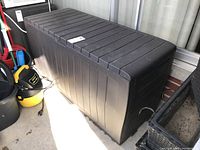 Closed view of large black rectangular resin outdoor storage bin with slatted lid, handle on front, and vertical grooves on sides.