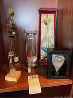 Photo showing the set of 5 timers and barometers including traditional hourglass, two cylindrical glass barometers, wall-mounted barometer, and black frame liquid barometer.
