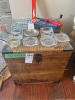 Front view of wood box with metal corners and latch, eight clear glass ashtrays and red rolling machine on top.