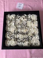 Front view of framed wall art with white artificial roses inside and text 'home sweet home' with date on glass.