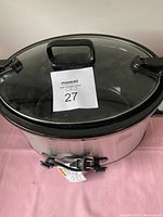 Oval slow cooker hot pot with clear glass lid, black handles, stainless steel exterior, and power cord placed on a pink surface against a white wall.