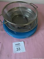 Photo of the stainless steel wire strainer with handles and blue round plate underneath on pink cloth, with lot tag visible.