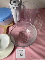 Photo showing clear glass bowl with floral pattern, white electric water pump, transparent plastic pitcher, blue-lidded plastic container, glass tumbler, ceramic plates, and wooden cutting board on pink surface.