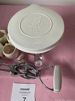 Top view of the white plastic cover with embossed Pampered Chef logo for the glass mixing bowl, rotary hand mixer and ceramic cups visible underneath on a pink surface.