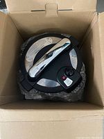 View of Living Basic electric pressure cooker inside cardboard box, showing stainless steel body, black handle and detachable lid with temperature control dial and red light indicator.