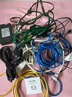 A tangled assortment of various cables including power cords, ethernet cables, USB cables, black power adapter, and a camera/webcam seen on a pink cloth background with a MaxSold number card.