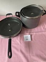 Photo of the pot and frying pan with lids on a pink cloth background showing handles and lid knobs.