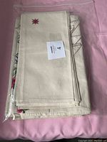 Folded fabric table covers stacked, showing plain cream fabric with red and blue floral embroidery in plastic bag.