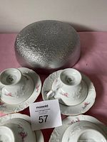Two white floral teacups and matching saucers with a textured silver bowl in the background.