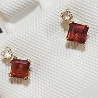 Close-up image showing the pair of garnet and diamond yellow gold earrings on a white textured background.