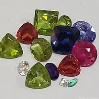 A group of 14 assorted genuine faceted gemstones varying in shape and color including greens, blues, purples, pinks, oranges, whites, and teal.