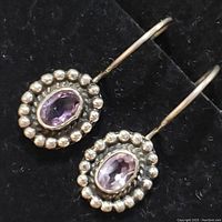 Close-up of two sterling silver earrings with central oval faceted purple amethyst stones surrounded by raised silver bead border, hanging on curved hooks. Placed on black background.