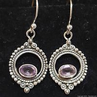 Front-facing photo of pair of sterling silver amethyst earrings displayed on black background showing circular designs with beaded silver details and central oval amethyst stones.