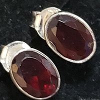 Close-up image showing two oval faceted garnet stones bezel set in sterling silver earrings on a black background.