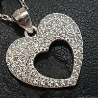 Close-up front view of the heart-shaped pendant showing pave-set small clear stones and the open heart cutout in the center on a dark background.