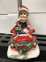 Front view showing girl with red Santa hat and present on car hood with wreath decoration on the front bumper.