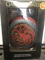 Photo of one Game of Thrones holiday ornament in original packaging. The ornament displays the Targaryen three-headed dragon in red and gold on a circular ornament inside a black box.
