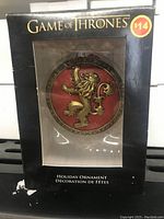 Game of Thrones holiday ornament in original black box packaging showing golden lion sigil on red background.
