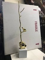 Full view of the golden tree branch sculpture with bird and hand base against a background box.