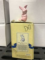 Photo of pink Piglet figurine sitting with arms crossed on yellow-striped box with Pooh branding.