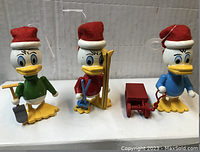 Three Huey Duck Christmas ornaments sealed in plastic with red Santa hats, each holding or pulling different accessories